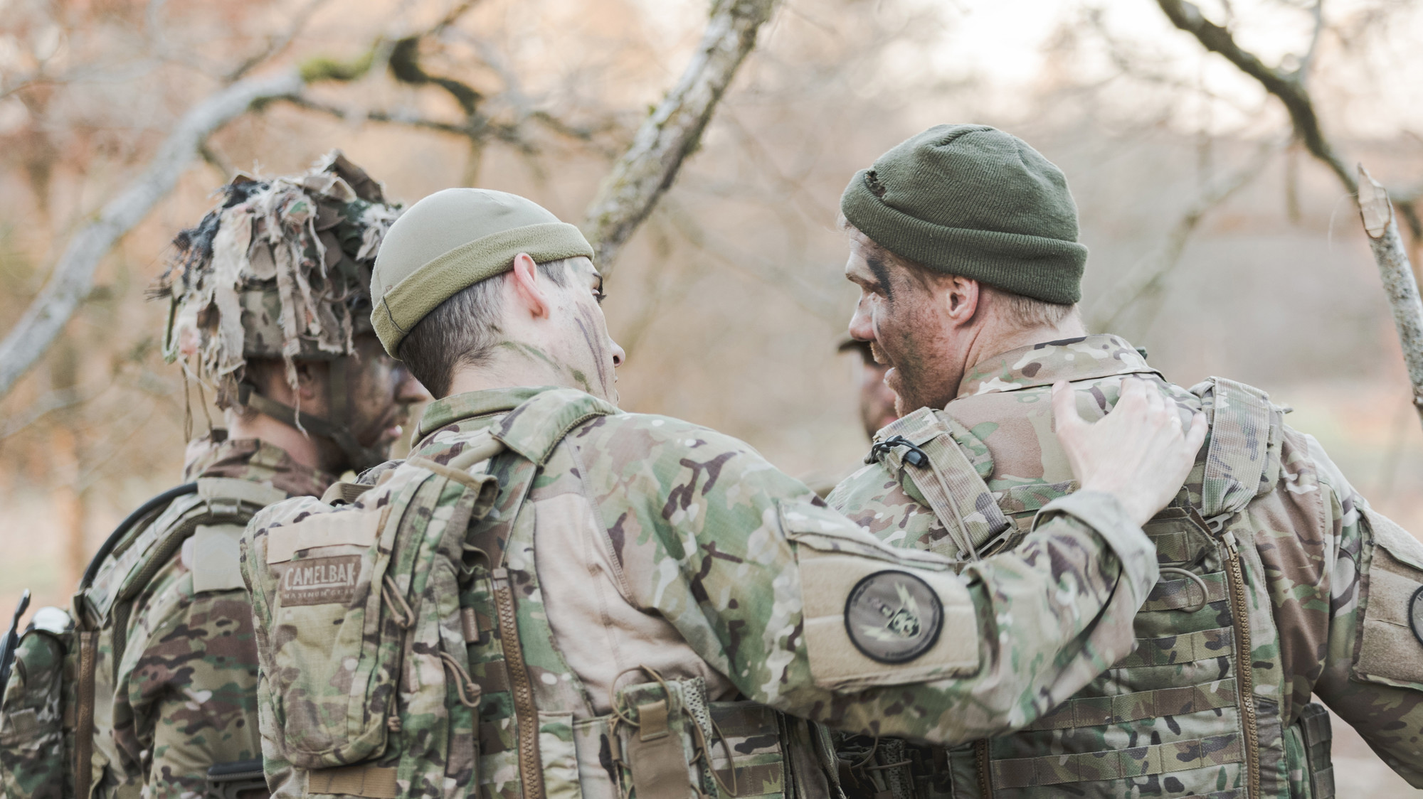 Tre soldater i camouflageuniform deler et kammeratligt øjeblik udendørs med træer i baggrunden. Stemningen udstråler sammenhold.