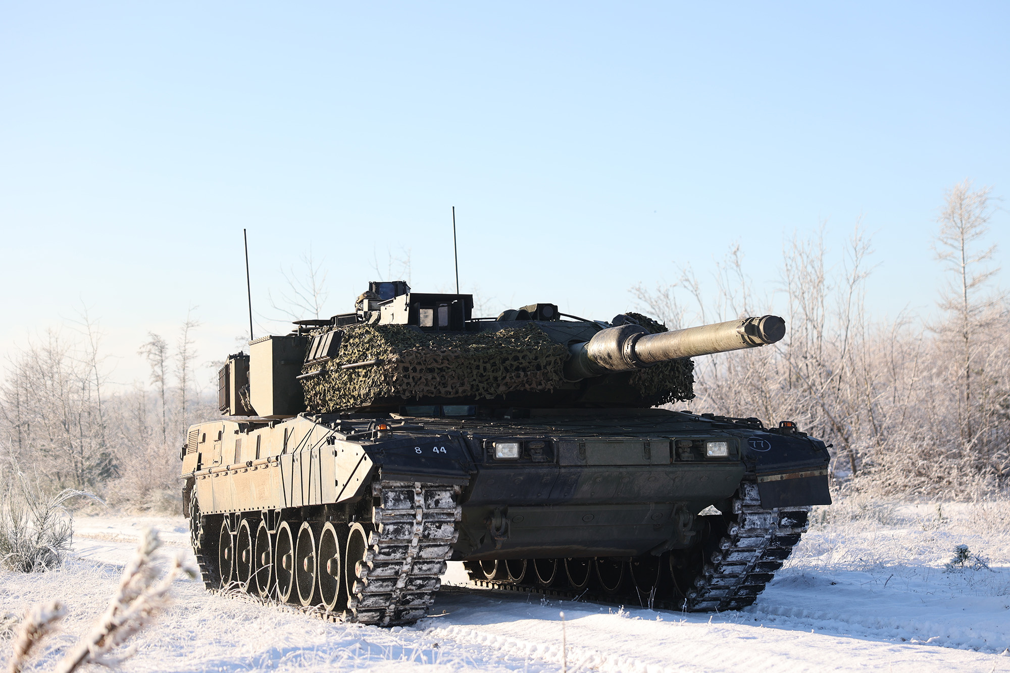 Militært kampvogn med camoufleret overflade og lang kanon på snedækket terræn omgivet af frostklædte træer.