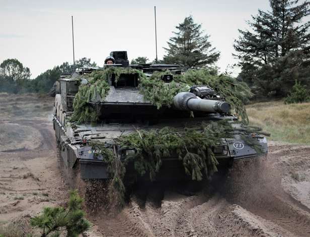 Kampvogn med camouflage af grene bevæger sig gennem sandet terræn, soldat ses delvist over tårnet, træer i baggrunden.