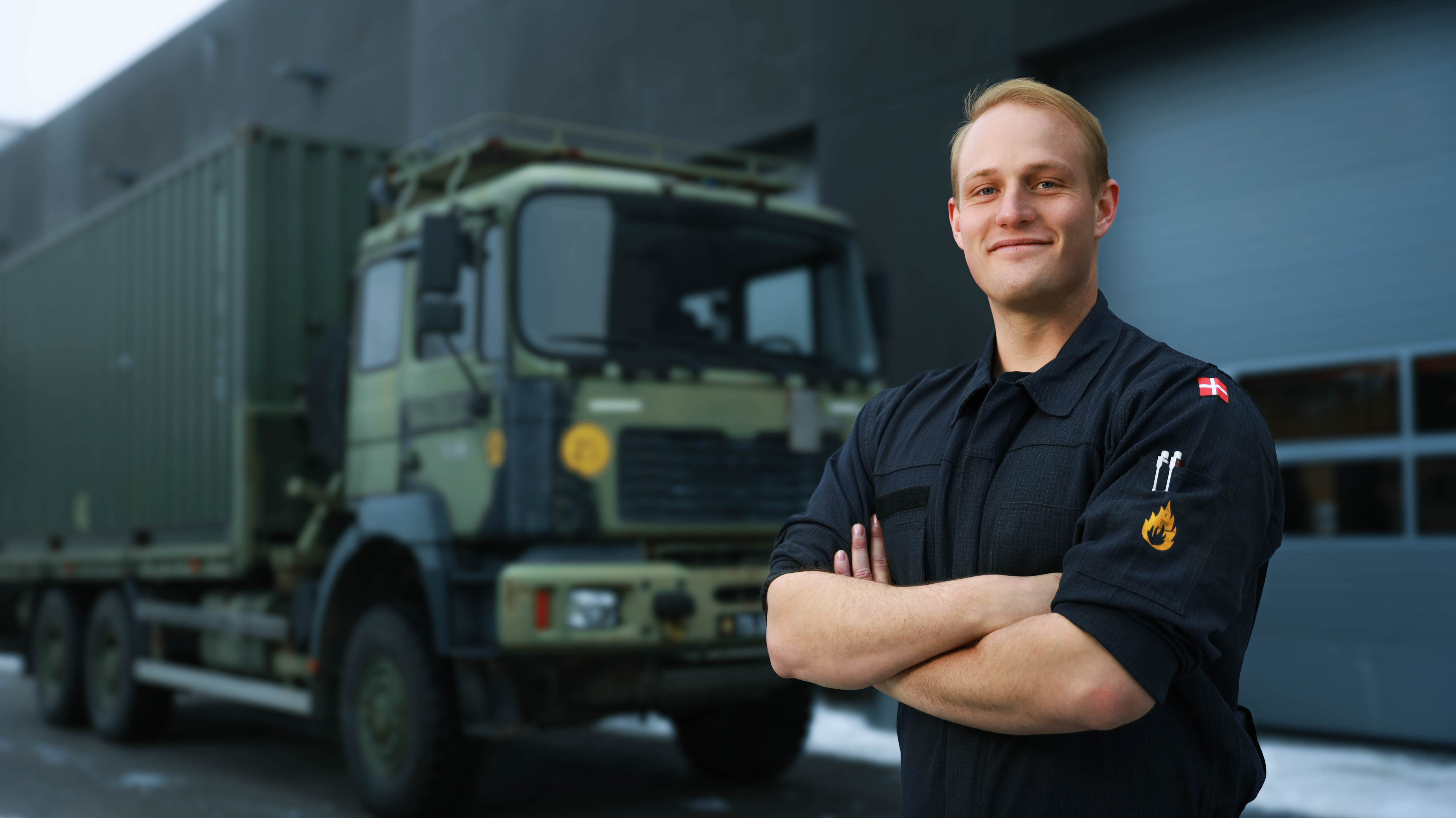 Mand i sort uniform med brandmands- og flagmærker står foran en camoufleret, militærgrøn lastbil i professionelt miljø.