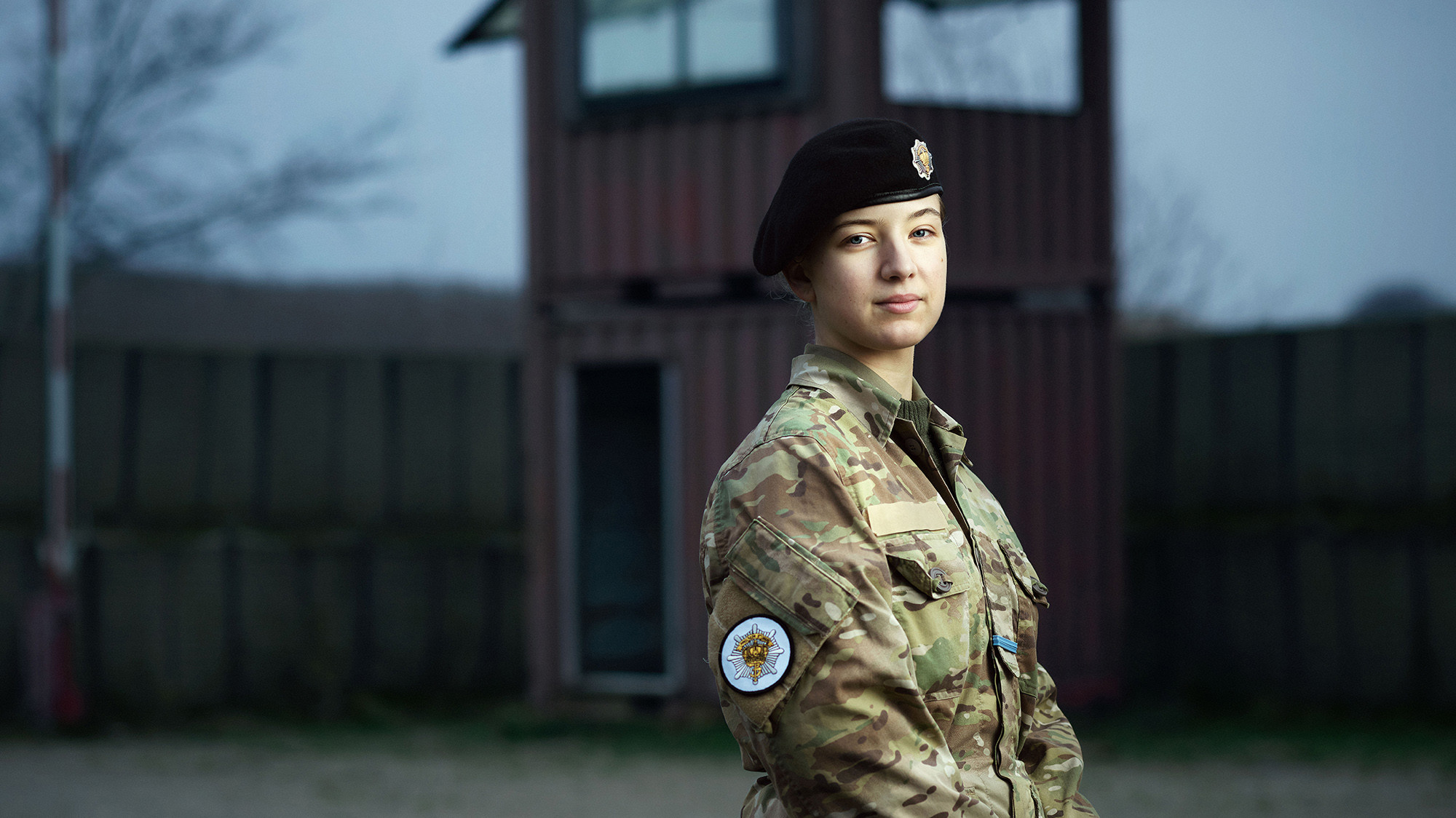 Kvinde i militæruniform og sort baret med badge smiler selvsikkert foran en vagtpost og hegn i dæmpet belysning.