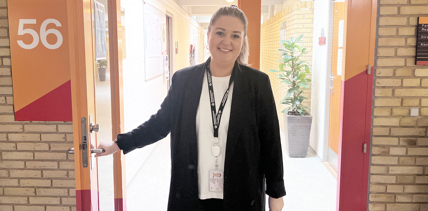 Kvinde i sort blazer og hvid skjorte står ved åben orange dør mærket "56" og byder velkommen i moderne gang med murstensvæg og plante.