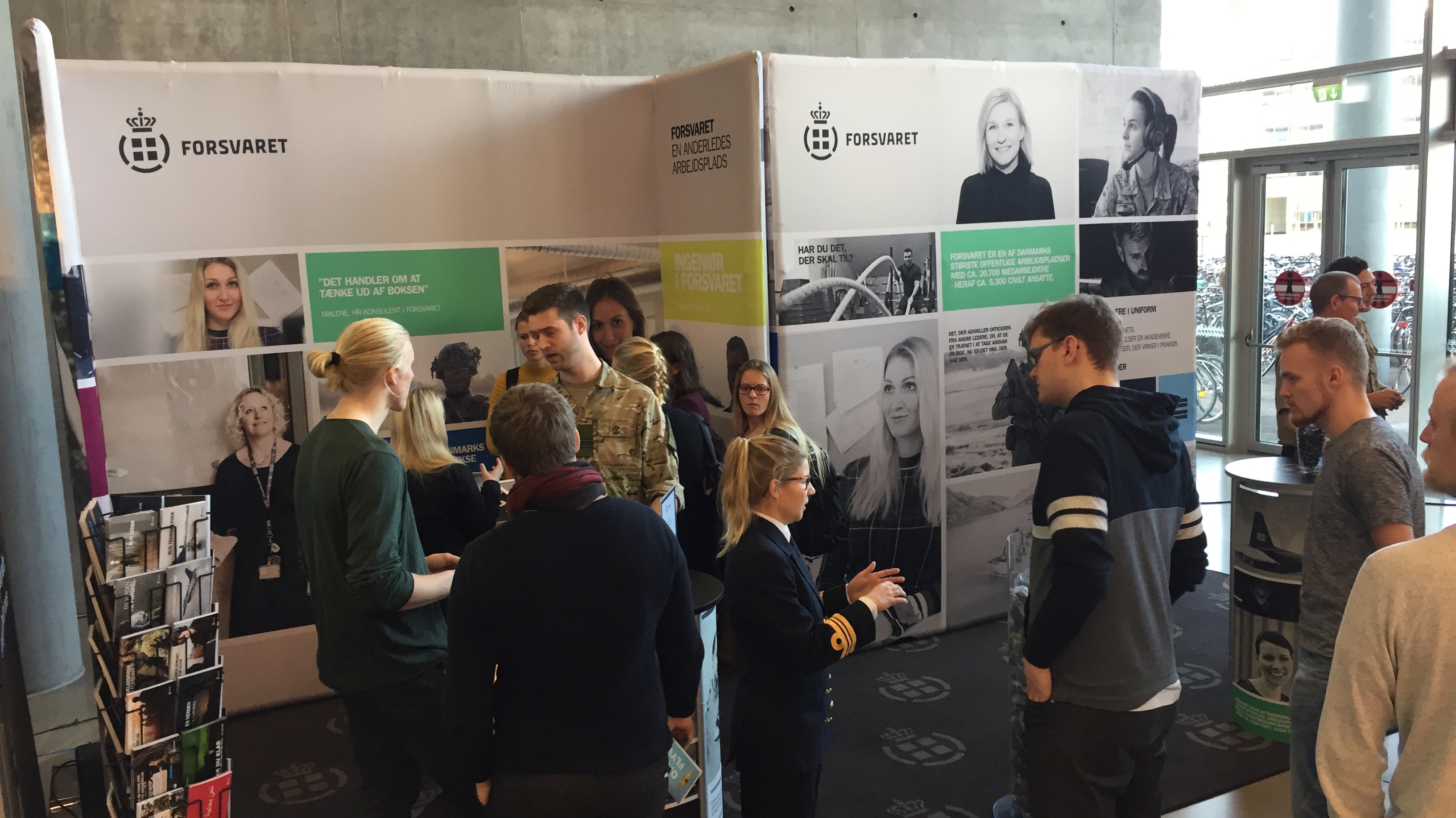 Rekrutteringsbod for Forsvaret med soldater i uniform og besøgende i dialog, omgivet af bannere om karrieremuligheder.
