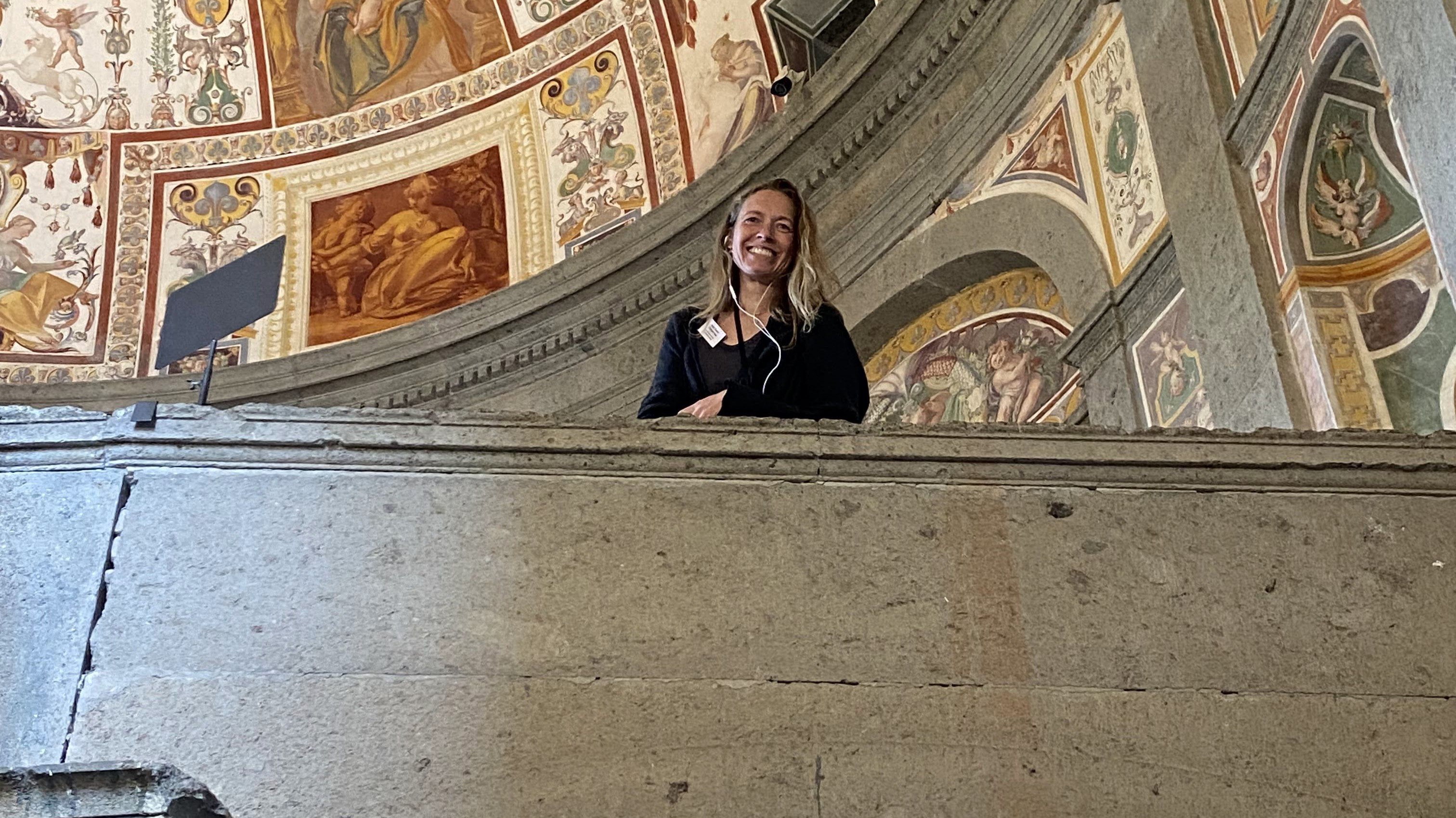 Kvinde med hovedtelefoner smiler på en stenbalkon foran dekorerede lofter og vægge i et historisk eller kunstnerisk rum.