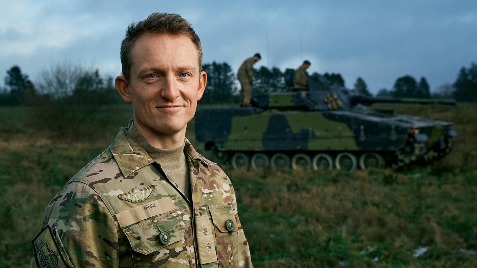 Soldat i camouflageuniform med insignier står selvsikkert foran en militærtank i et naturligt landskab under skyet himmel.