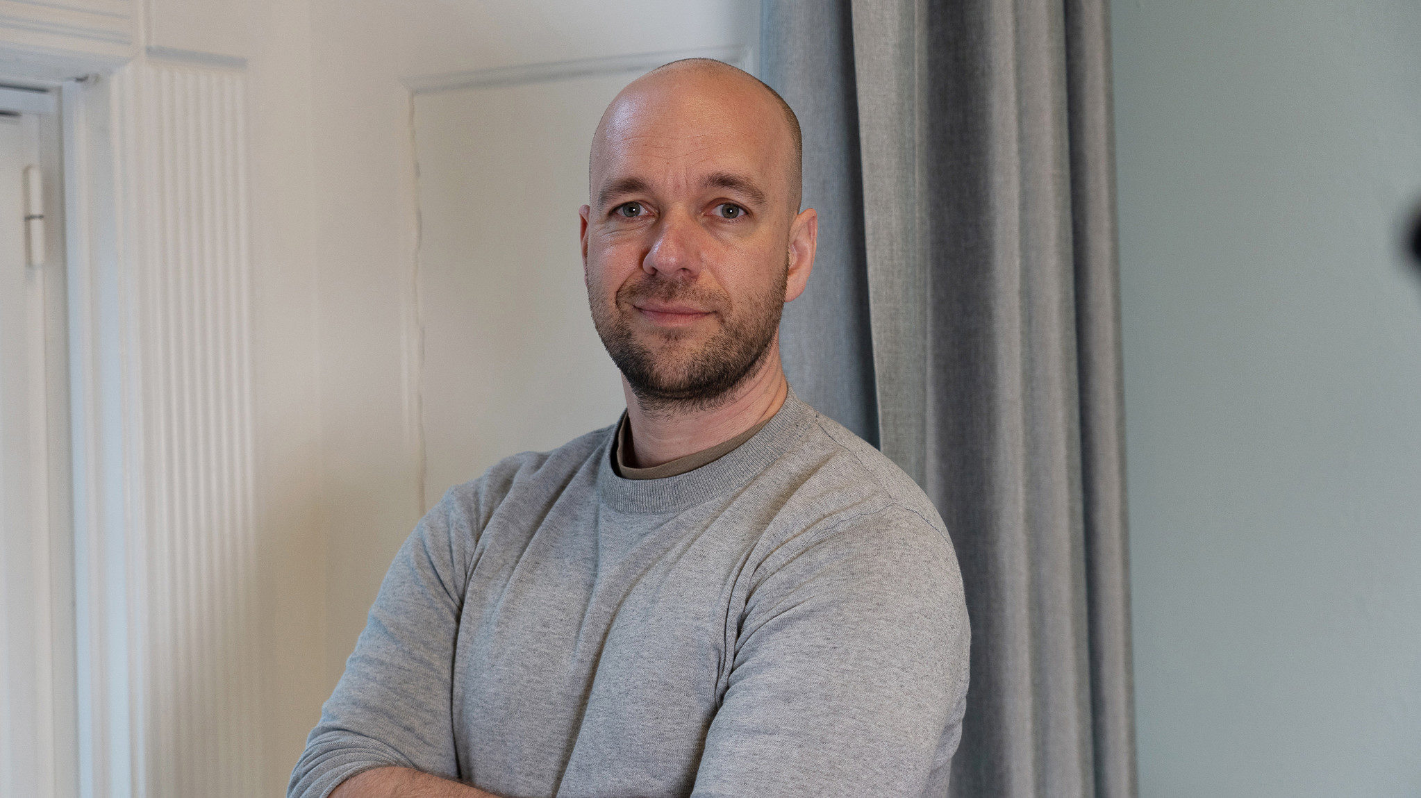 Mand med barberet hoved og kort skæg i lysegrå sweatshirt står afslappet med korslagte arme foran lys væg og gardin.