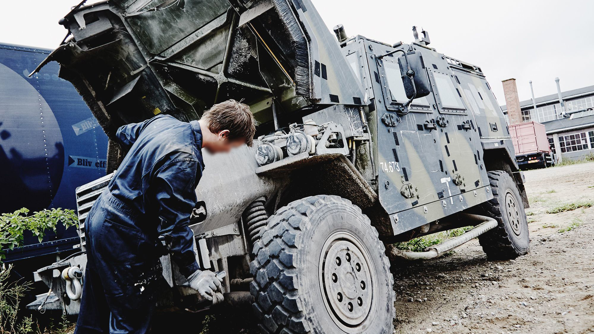 En pansret mandskabsvogn med åben bagende serviceres af en tekniker i mørk kedeldragt i et værkstedsmiljø.