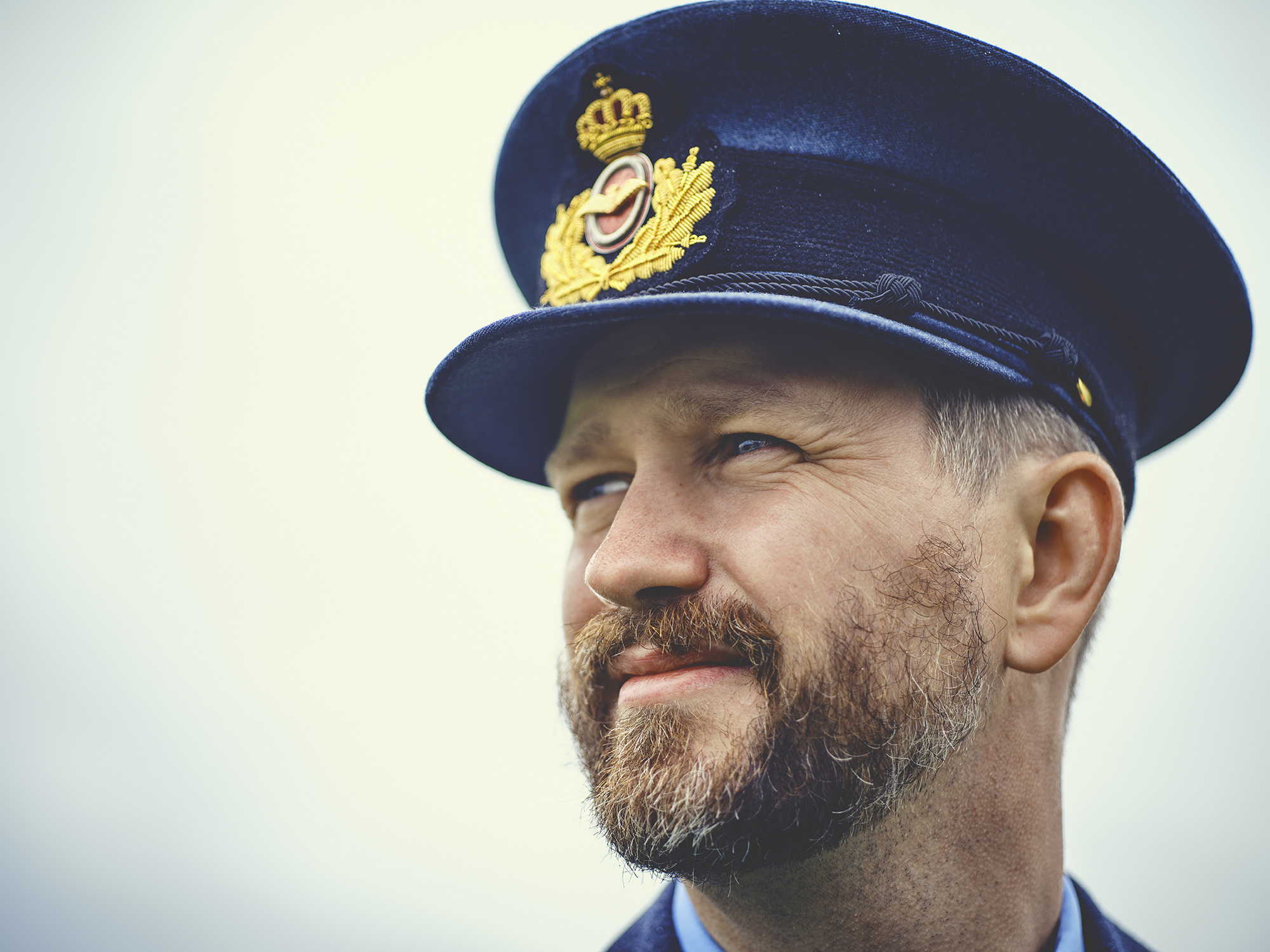 Mand i militæruniform med mørkeblå kasket med emblem, skæg og let smil. Fokus på ansigt og blå-grå farvetema.