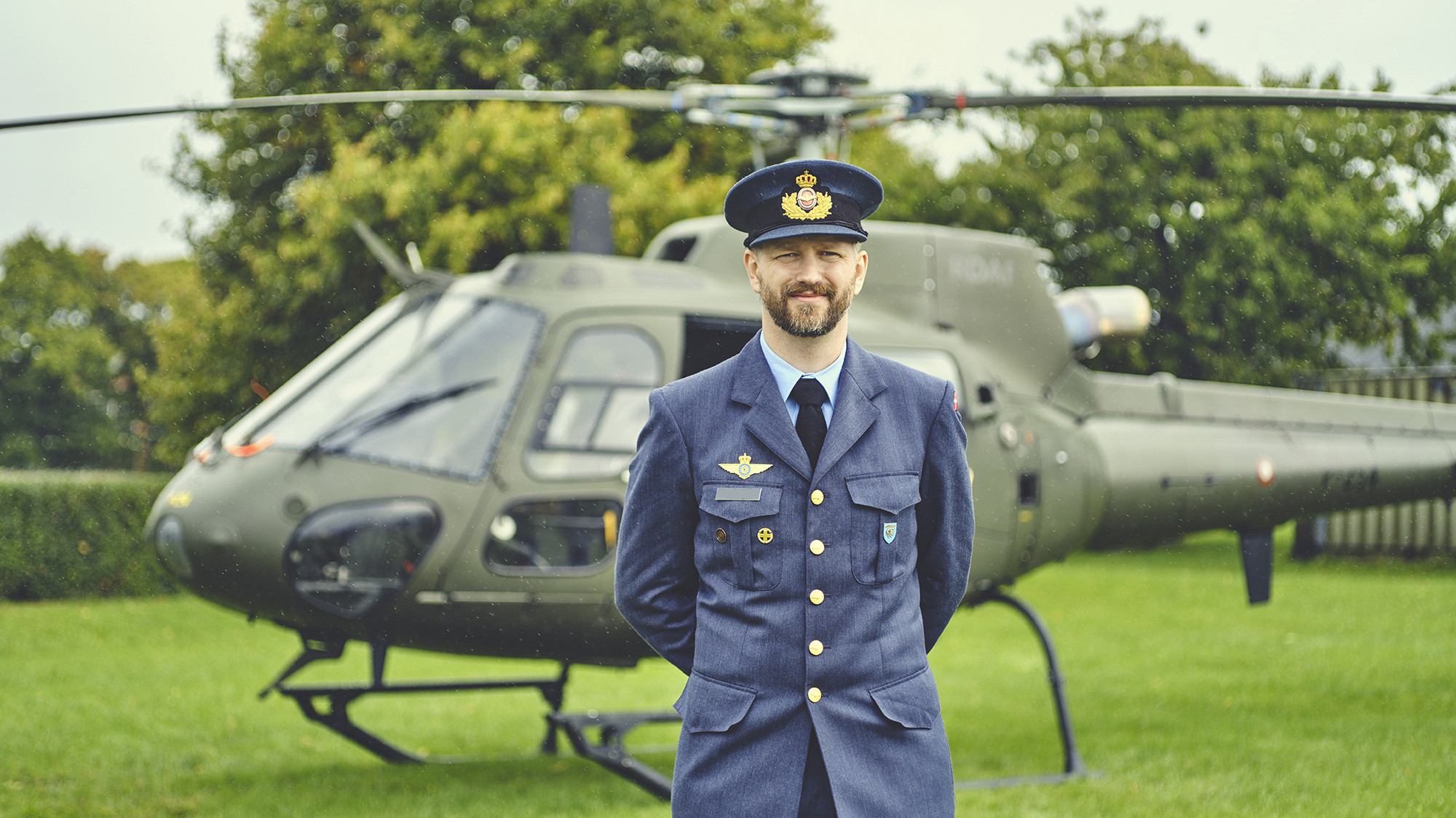 En mand i militæruniform med skæg og kasket står selvsikkert foran et camouflagehelikopter på en grøn mark.