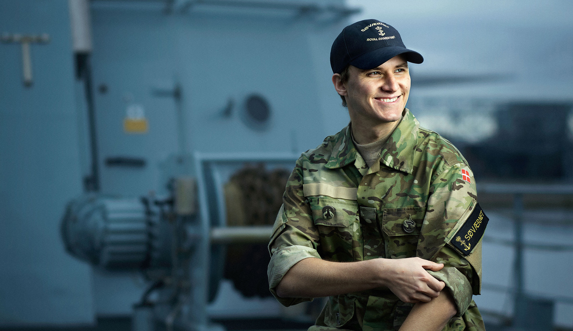 Ung mand i militæruniform med camouflagejakke og kasket med "Royal Danish Navy" sidder smilende på et skib.