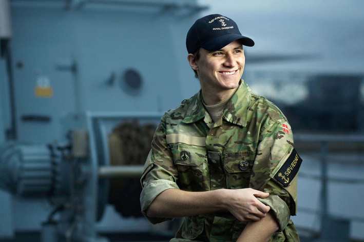 Ung mand i militæruniform med camouflagejakke og kasket med "Royal Danish Navy" sidder smilende på et skib.