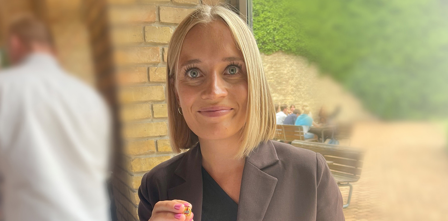 En kvinde med kort blondt hår iført sort top og mørk blazer smiler og holder en lille genstand, muligvis et smykke eller en dekorativ ting, op mod kameraet; baggrunden består af en murstensvæg og antyder personer, der sidder i en afslappet udendørs atmosfære med en varm og indbydende stemning.