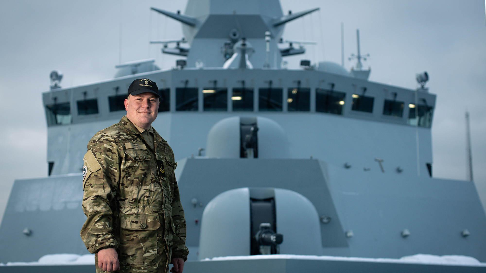 Soldat i camouflageuniform og sort kasket smiler foran moderne flådeskib med sne i baggrunden, hvilket udstråler stolthed og professionalisme.