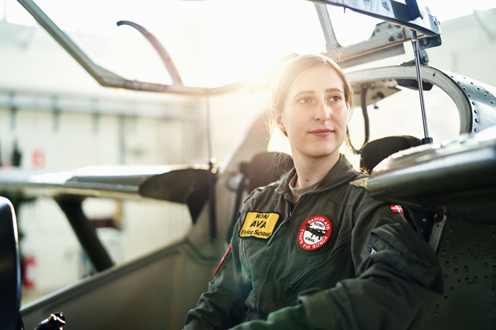 Ung kvindelig pilot i cockpit iført flyverdragt med patches fra Flyvevåbnet og en flyveskole. Sollys giver varm stemning.