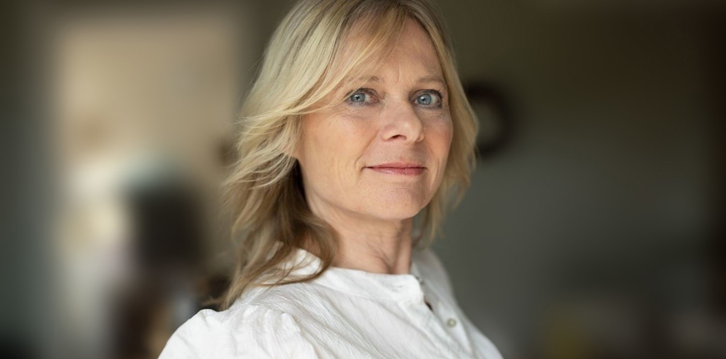 Kvinde med skulderlangt blond hår og hvid bluse ser roligt ind i kameraet med klare blå øjne, sløret baggrund.