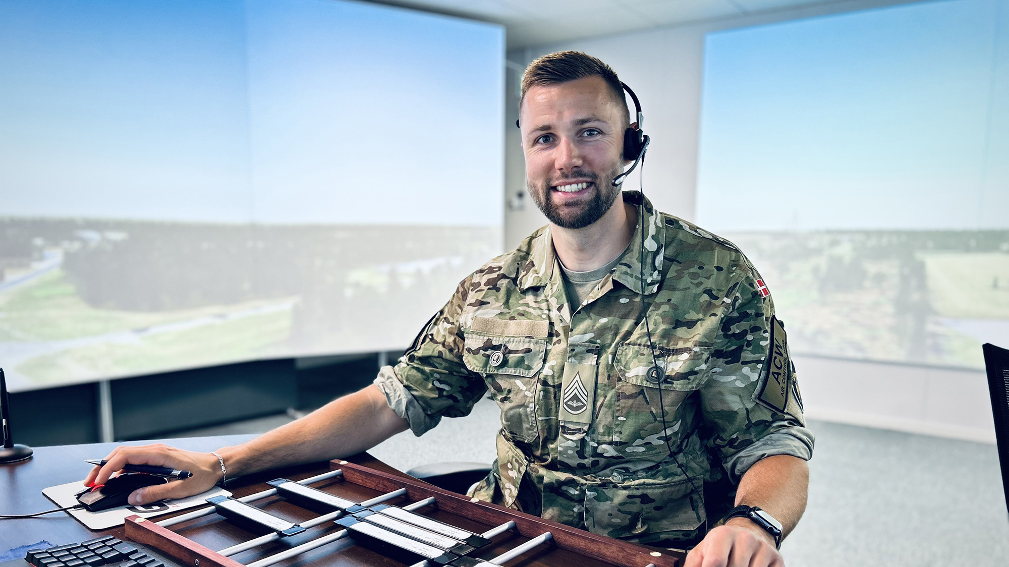 Soldat i camouflageuniform og headset smiler ved skrivebord med computerskærme, der viser et landskab i baggrunden.
