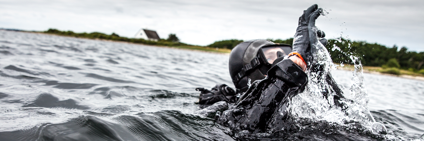 Dykker i våddragt med hænderne hævet og sprøjtende vand, iført hjelm og handsker, foran en kyst med skyer i baggrunden.