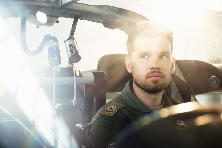 Ung mandlig pilot i cockpit med koncentreret blik, blødt sollys i baggrunden og to sorte mikrofoner over hovedet.