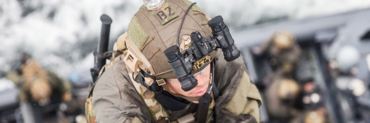 Soldat i taktisk udstyr med natkikkerter og pandelampe, liggende i maritimt miljø sammen med hold, klar til specialoperation.