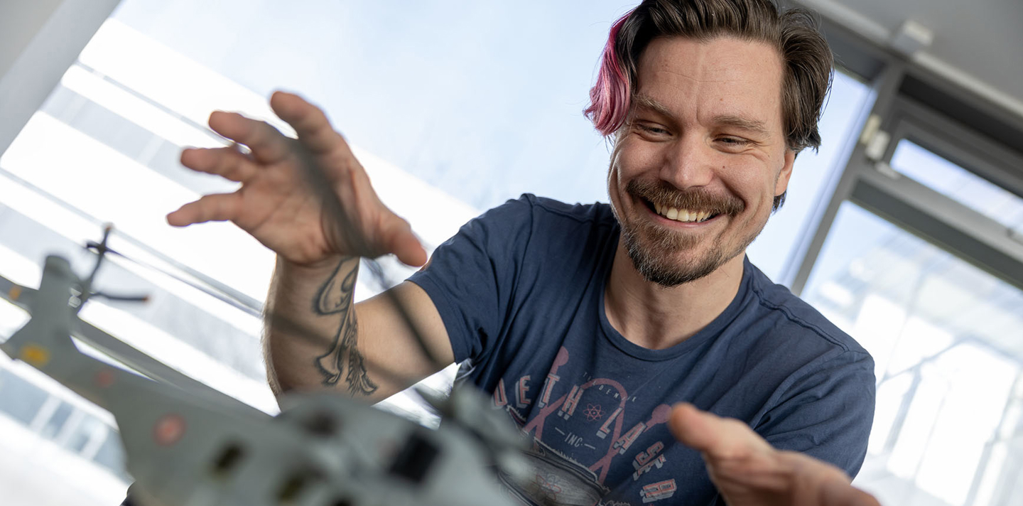 Mand med kort hår og tatoveringer på armen leger begejstret med en modelhelikopter ved et bord i et lyst, moderne rum.