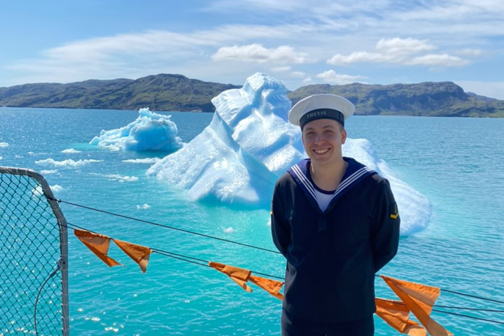 Ung mand i marineuniform står på skibsdæk med isbjerge i blåt vand og bjerge i baggrunden. Hans hat har navnet "THETIS."