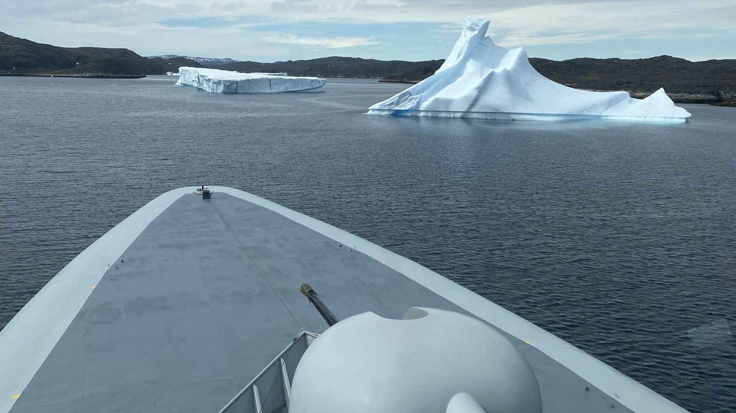 Udsigt fra et skibs forstavn over vand med store isbjerge i det fjerne, dækket er tydeligt i forgrunden under en skyet himmel.