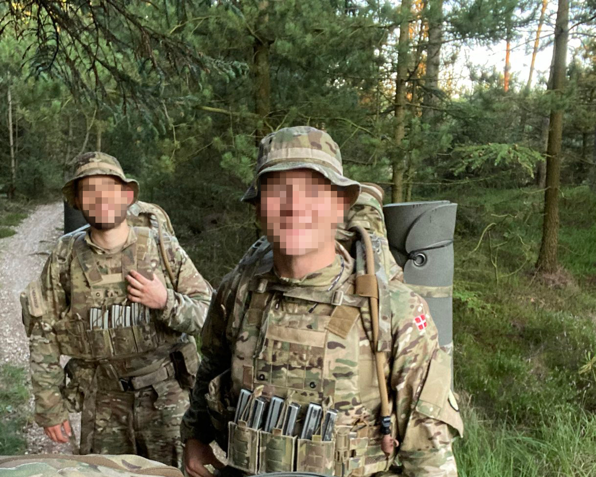 To personer i militær camouflagetøj og taktisk udstyr står i en skov, begge med hatte, én med rygsæk. Baggrunden er sløret.