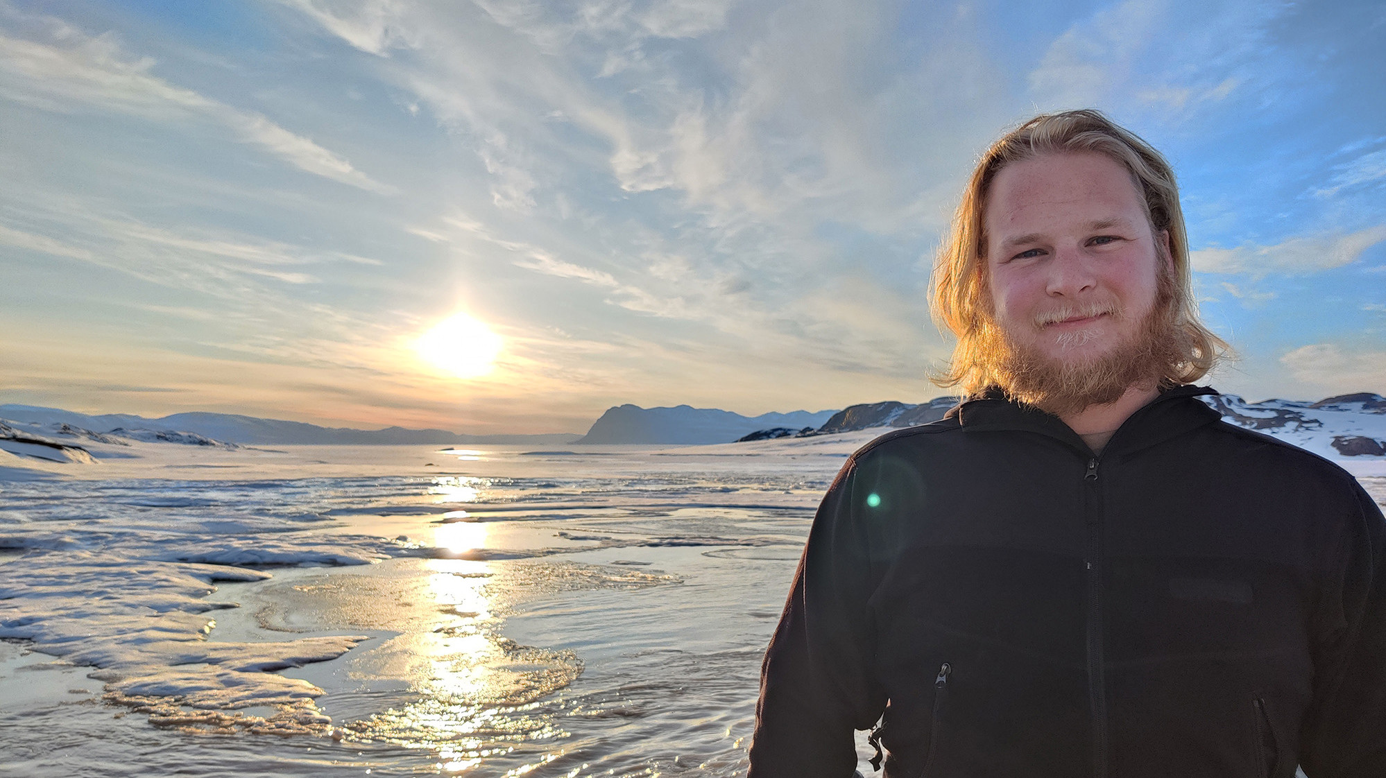Ung mand med langt hår og skæg smiler i et snedækket landskab med solnedgang, vand, is og bjerge i baggrunden.