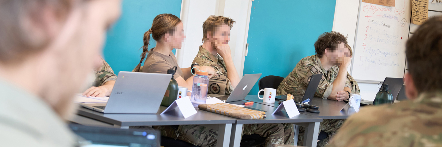 Flere personer i militæruniformer sidder rundt om et bord med bærbare computere og krus, foran en blå væg og whiteboard.