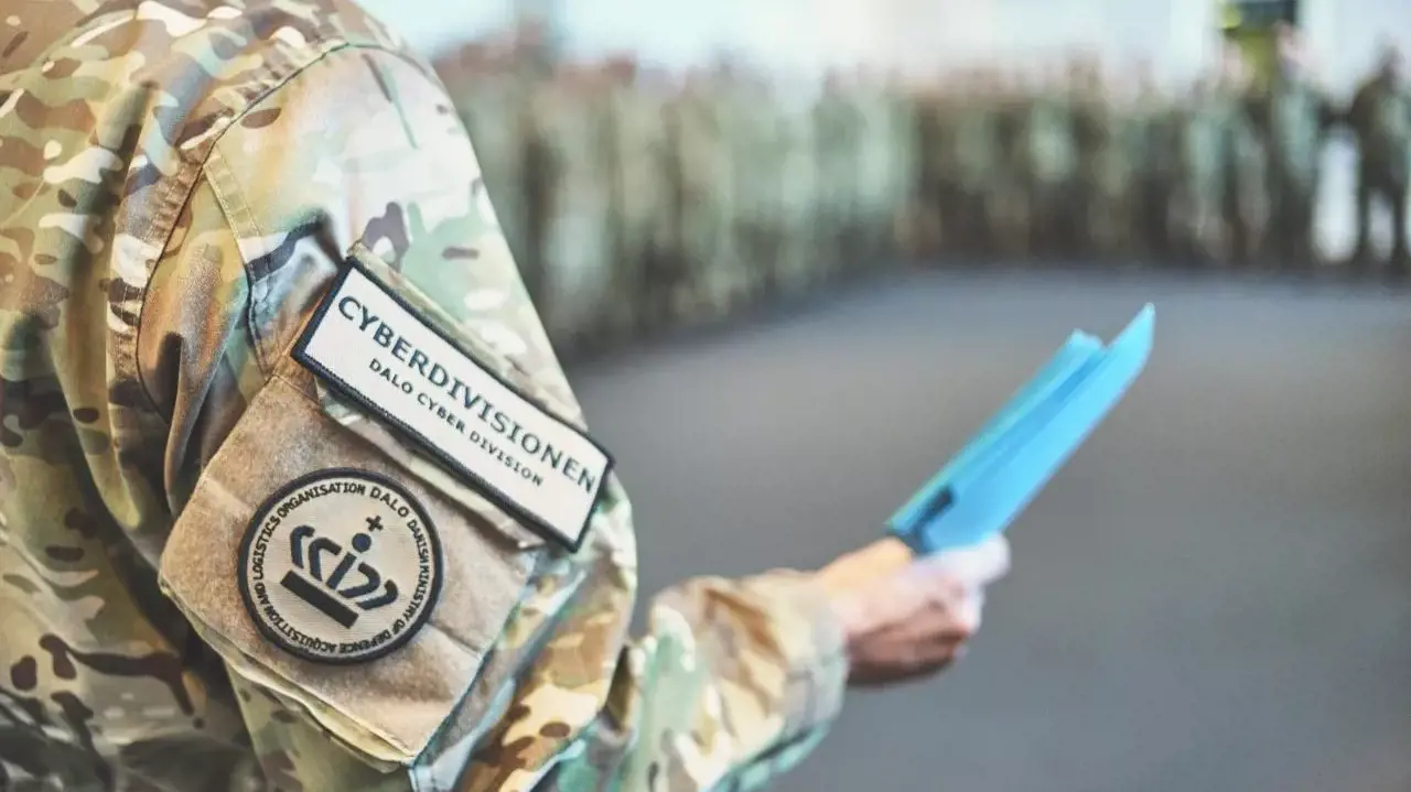Militærperson iført camouflageuniform med CYBERDIVISIONEN-mærke holder et blåt dokument; sløret militærgruppe i baggrunden.