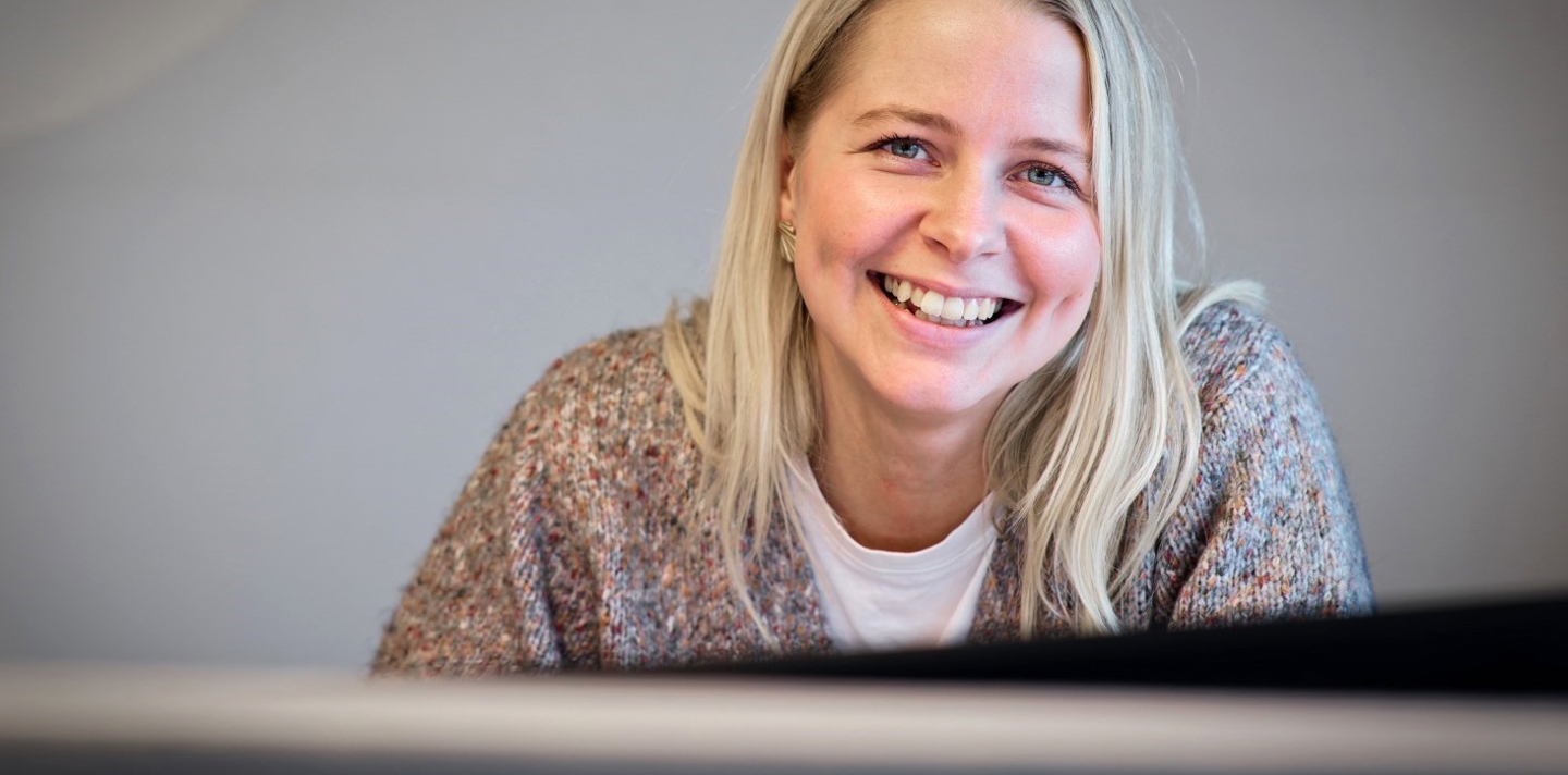 Smilende kvinde med langt lyst hår i farverig sweater sidder ved et skrivebord, med fokus på hendes glade udtryk og elegante øreringe.