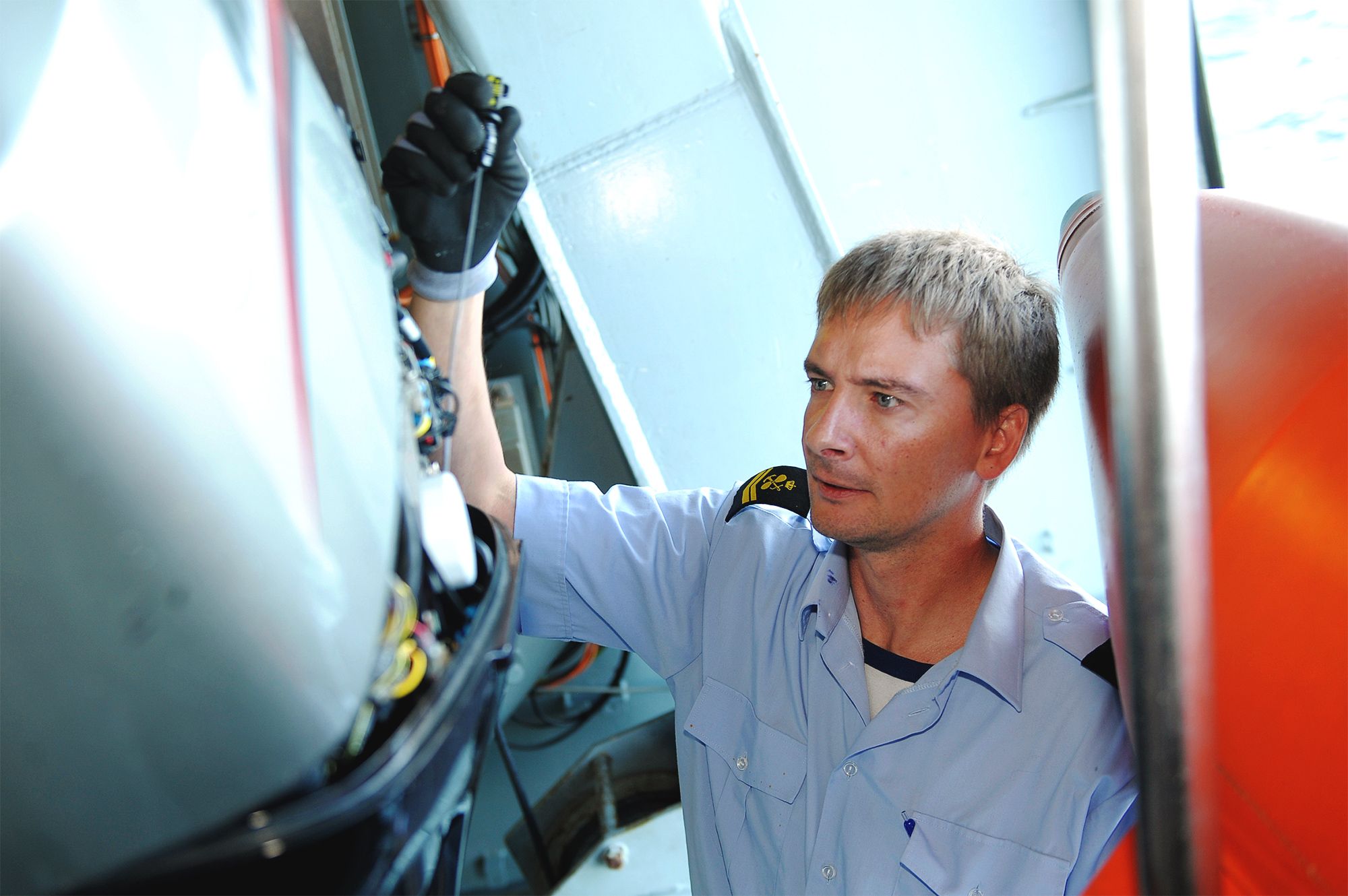 En tekniker i lyseblå uniform med skulderinsignier arbejder fokuseret med udstyr ombord på et skib med synlig redningsflåde.