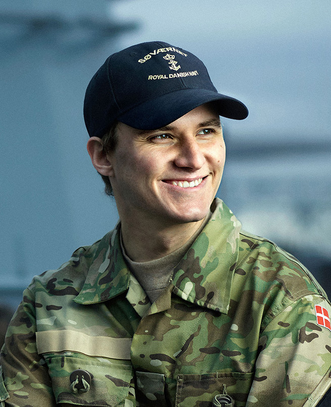 Ung mand i camoufleret uniform og marinekasket med teksten “SOVERÆNIT” og “ROYAL DANSK MARINE”, smilende udendørs.
