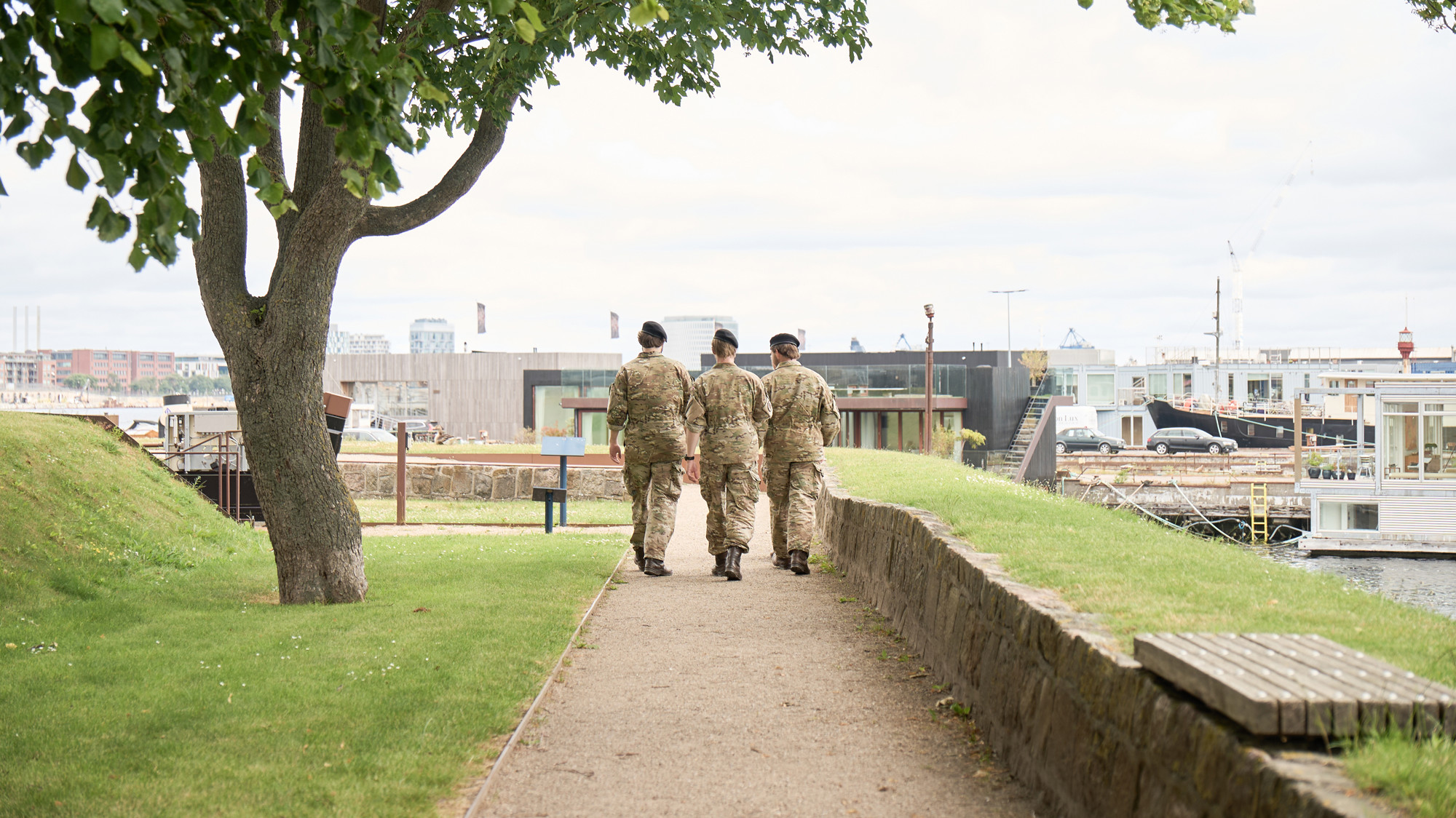 Tre personer i militæruniformer går på en sti med grønne omgivelser og moderne bygninger ved vandet i baggrunden.
