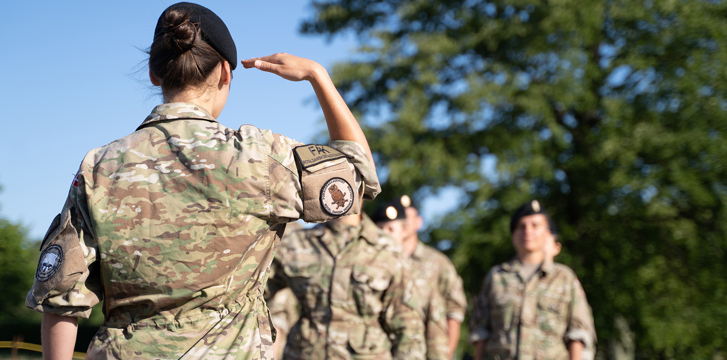 Kvindelig soldat i uniform med ryggen til, gør honnør for en gruppe soldater