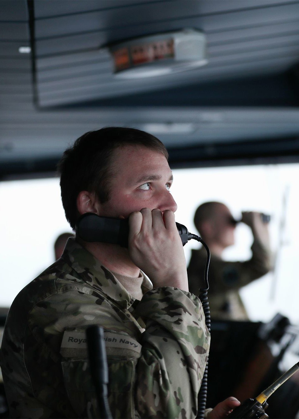 Servicemand i camouflageuniform taler i telefon ombord på et skib, mens kollega overvåger omgivelserne med kikkerter.