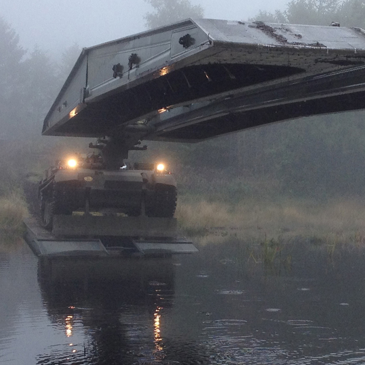 Militær bro-lægningskøretøj med hævet brosektion over vand, omgivet af tæt tåge og reflekterende vandoverflade.
