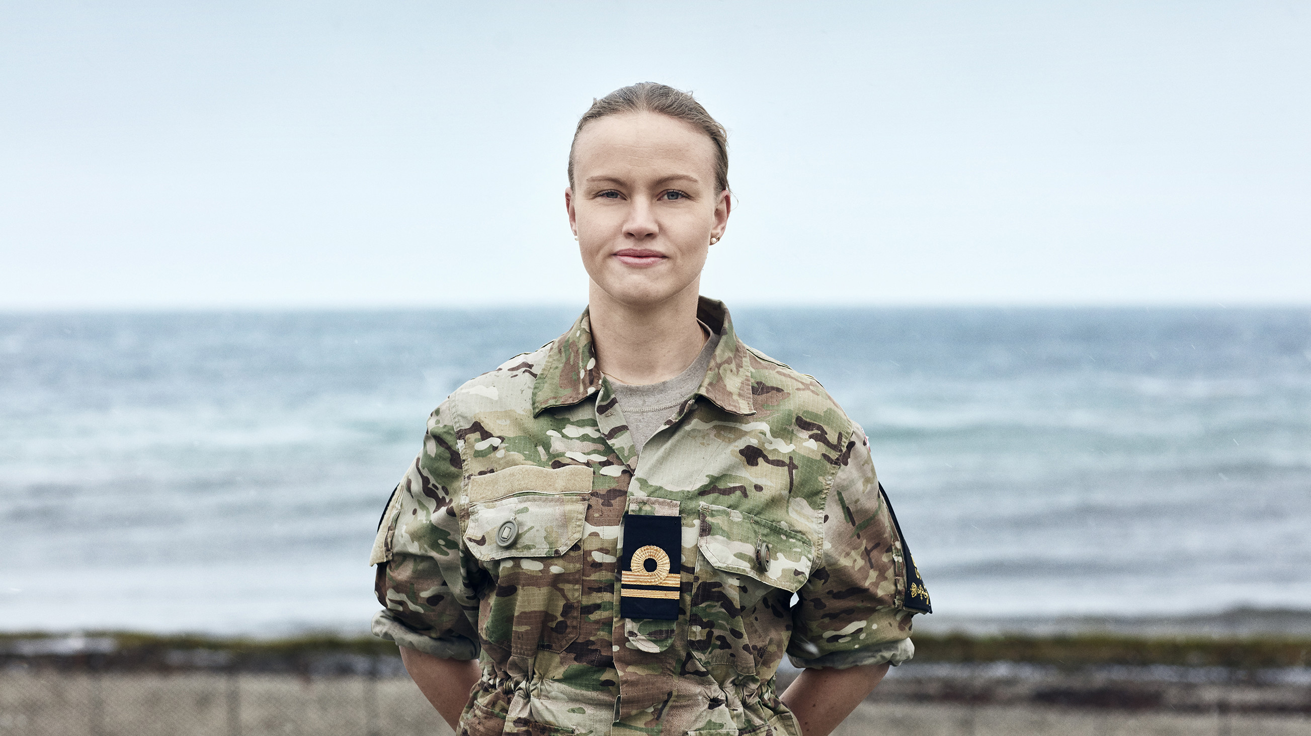 Kvinde i militæruniform fra Søværnet står foran havet og kigger roligt mod kameraet.