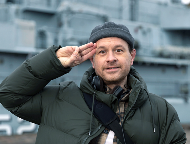 Christian Fuhlendorff i vinterjakke og hue giver et uformelt salut foran et stort gråt skib.