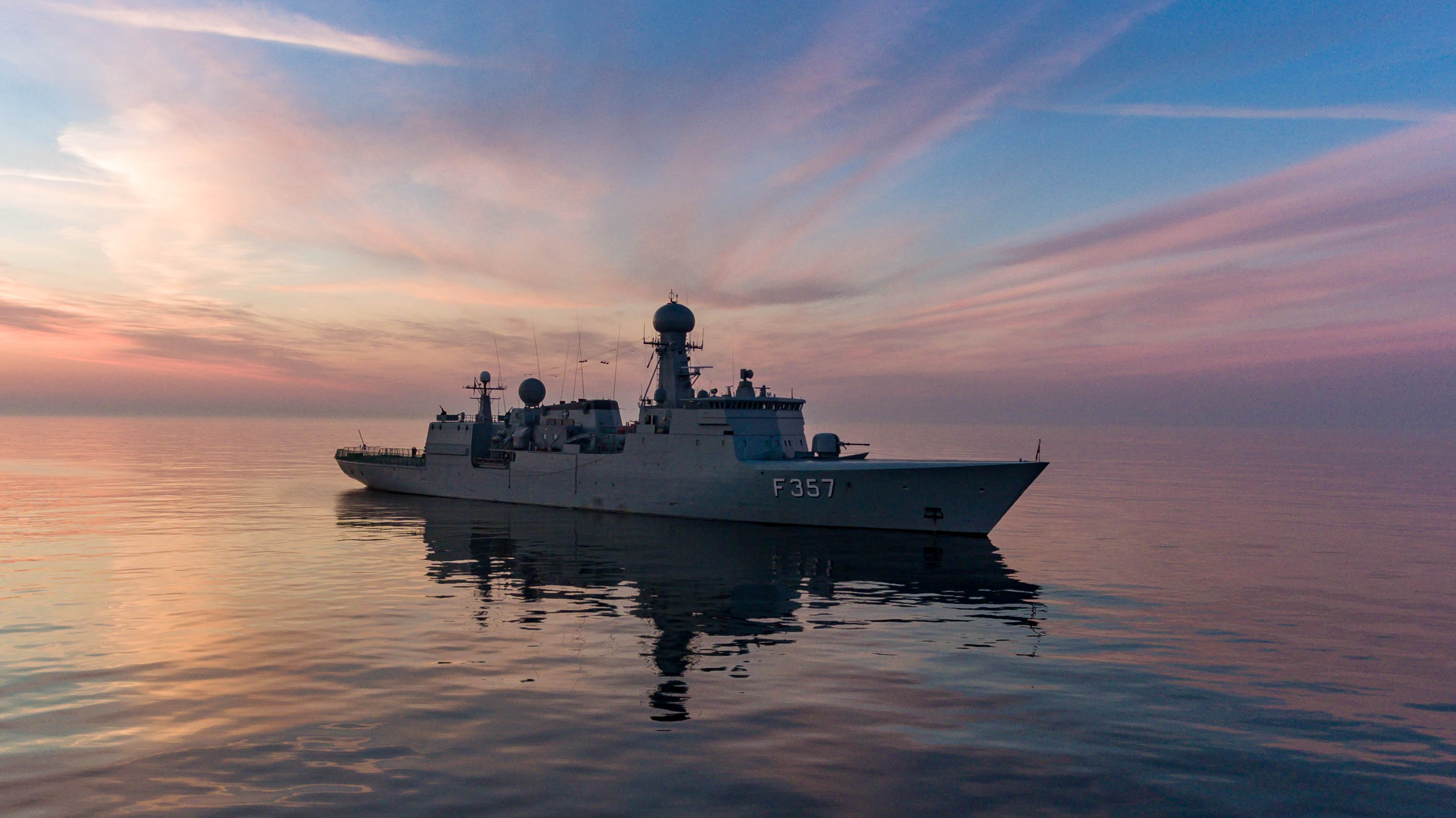 Et flådefartøj ligger stille på roligt vand i solnedgangen. Himlen er rosa og blå, og skibet spejler sig i havet.