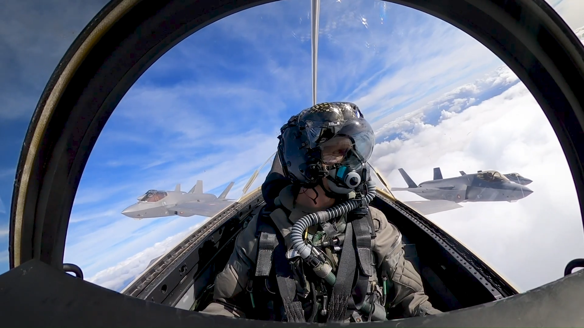 Pilot i cockpit med udsigt til to F-35-fly, der flyver i formation over skyerne.