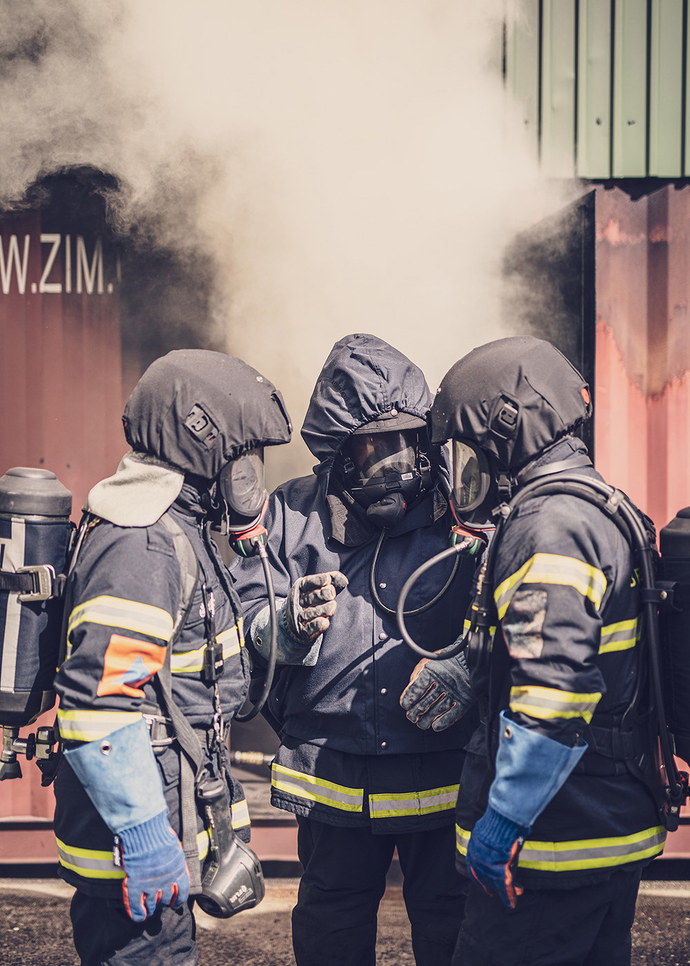 Tre brandmænd i fuldt røgdykkerudstyr holder taktikmøde foran en container med tæt røg under brandslukningsøvelse.
