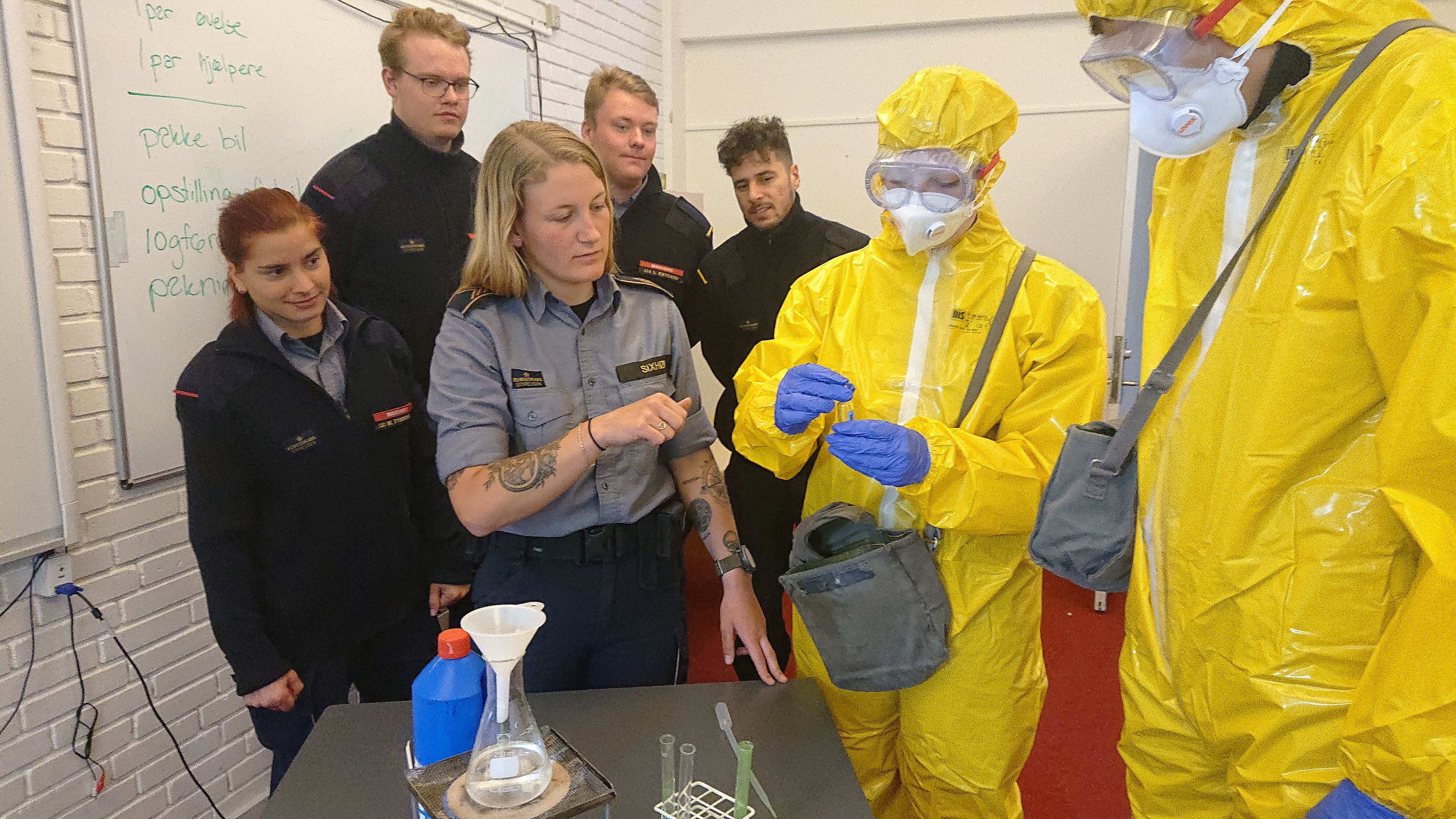 Instruktør forklarer kemikaliehåndtering til kursister i beredskabsuddannelse, mens to deltagere i gule hazmat-dragter håndterer en prøve.