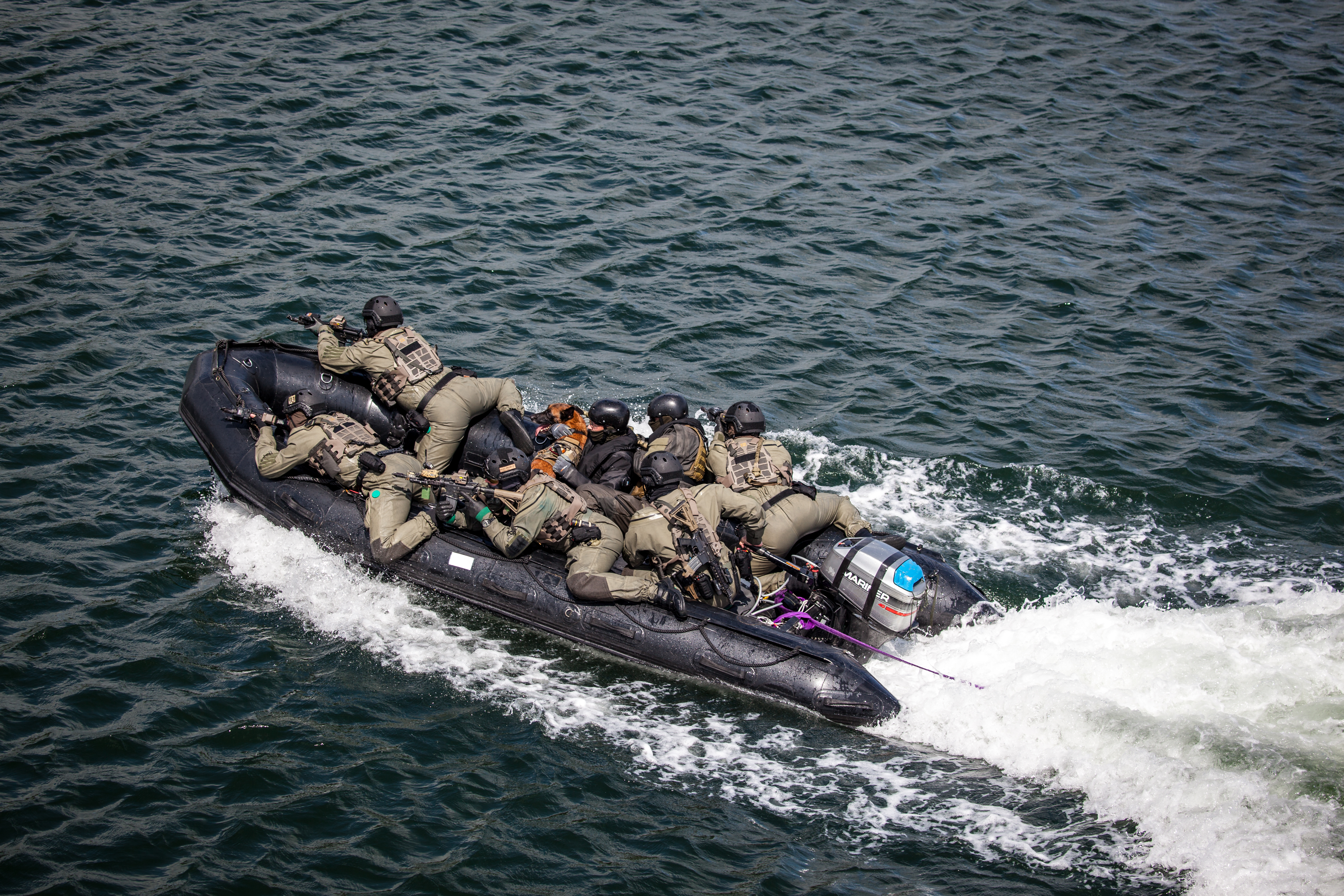 Bevæbnede soldater i taktisk gear ligger fladt i en sort gummibåd under høj fart på åbent hav.