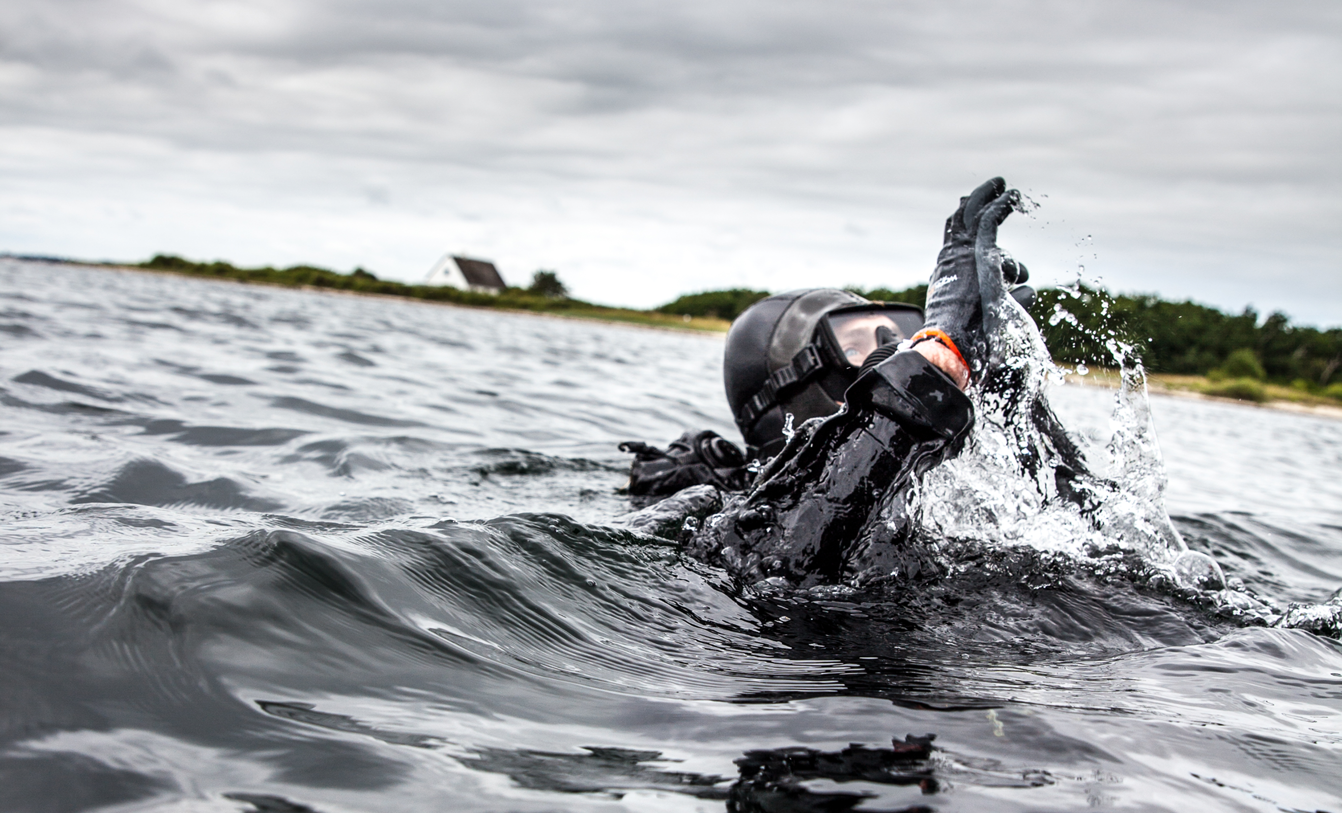 Dykker iført sort taktisk udstyr og hjelm bryder vandoverfladen tæt på kysten under en operation.