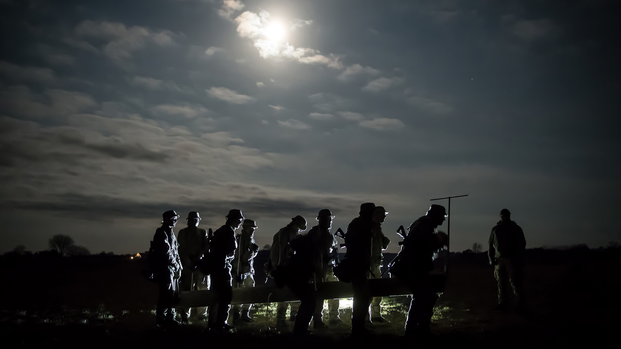 Silhuetter af soldater med udstyr i måneskin under en natlig militærøvelse i åbent terræn.