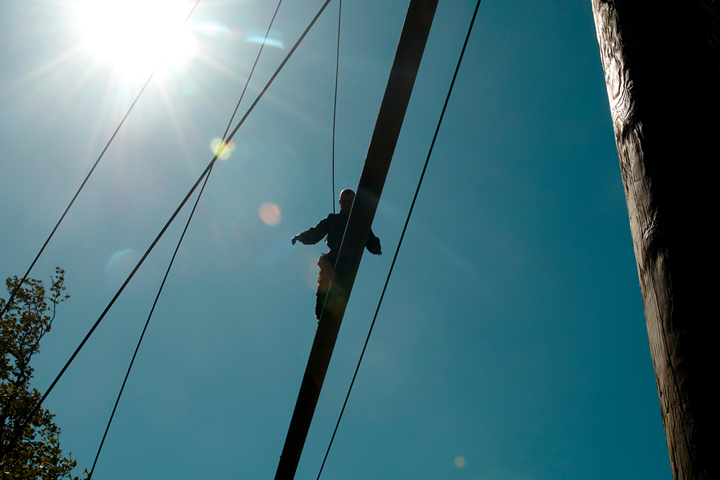 Person balancerer højt på en træbjælke i en forhindringsbane med sikkerhedsliner, mens solen skinner klart.