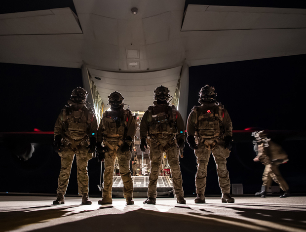 Fem soldater i fuld kampuniform står opstillet foran et militærflys åbne lastrampe om natten, klar til indsættelse.