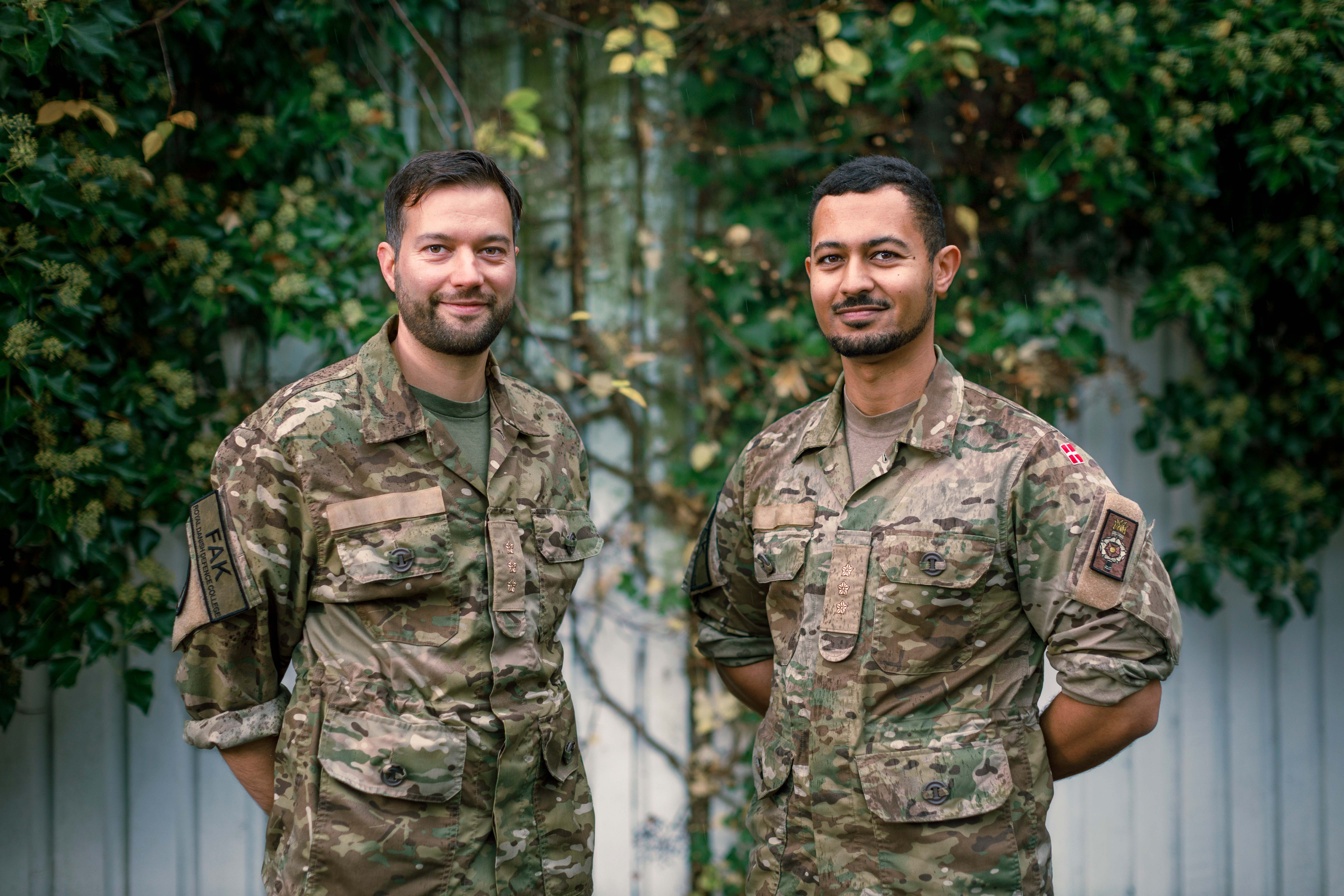 To danske sprogofficerer i uniform står smilende foran en baggrund af grønne planter og et hvidt hegn