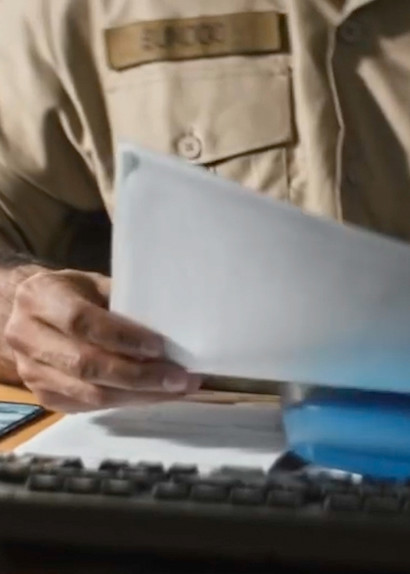 En person i uniform sidder ved et skrivebord og gennemgår dokumenter, mens hænderne bladrer i papirer foran en computer.